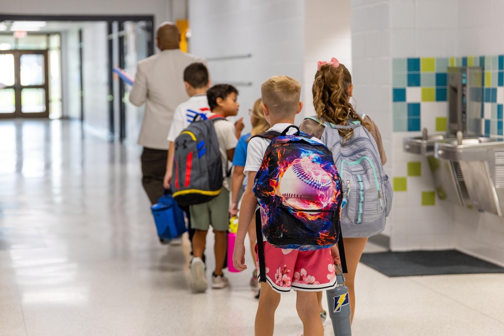 Children wearing backpacks walk down a school hallway with a man in the background. Multicolored tiles line the wall.