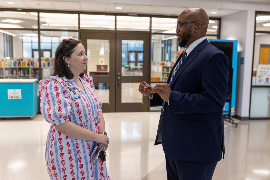 A man and a woman stand and talk in a school hallway. Behind them are doors, shelves, and a blue container.