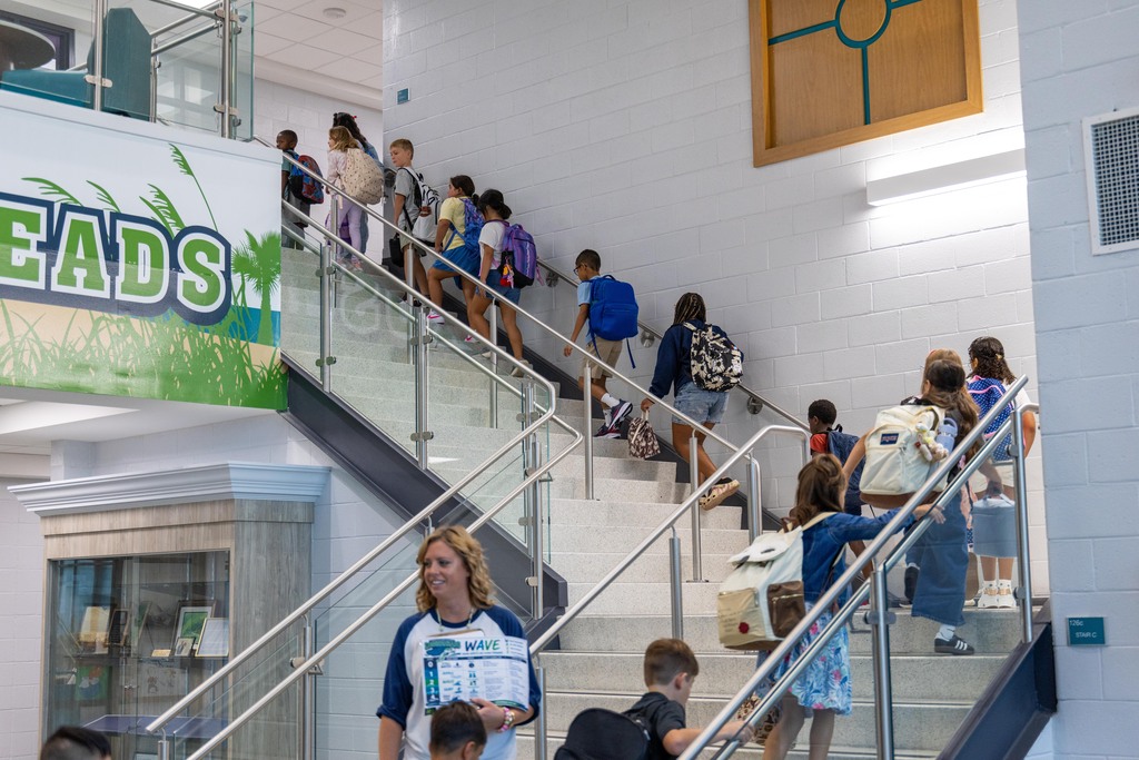 A group of children in school uniforms and backpacks climb stairs in a school building. A sign reads "HEADS."