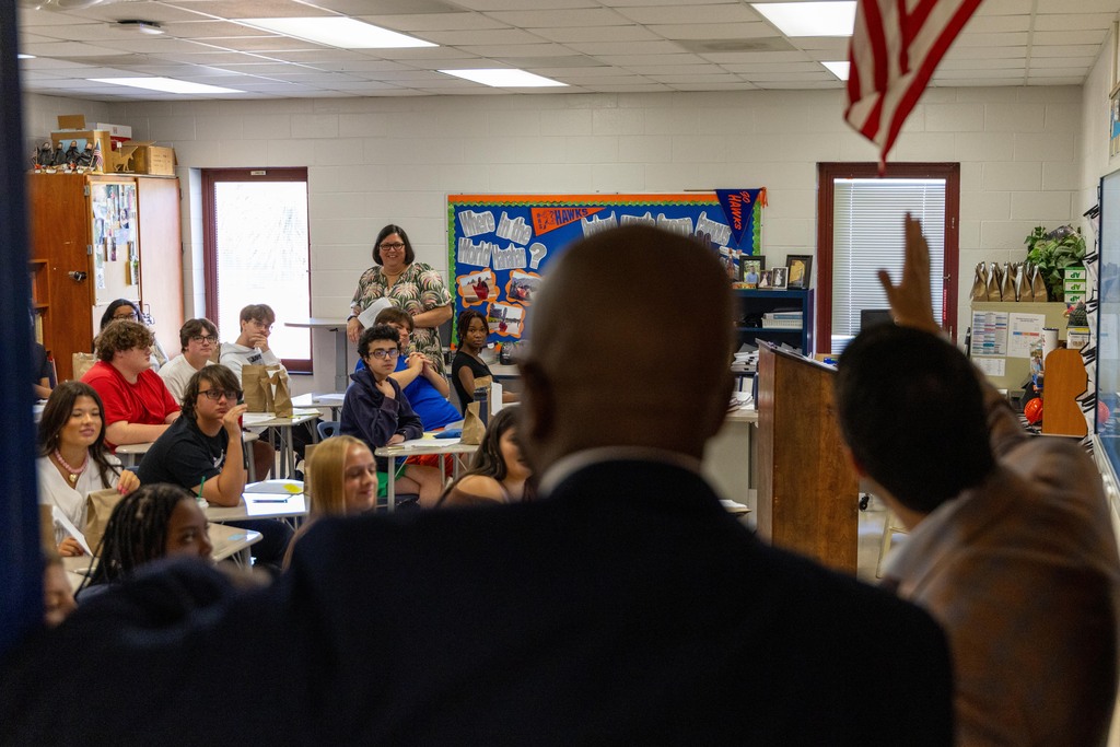 A group of people, including an adult man and woman, are sitting in chairs in a classroom.