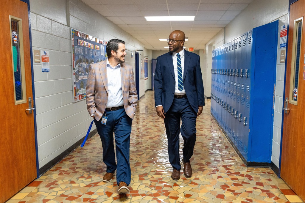 Two men in business attire walk down a hallway, past lockers. One wears a light-colored suit, the other a dark suit.
