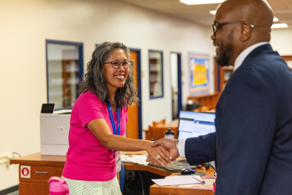 Two individuals shake hands in an office setting. The woman wears a pink shirt and glasses, the man a blue suit.