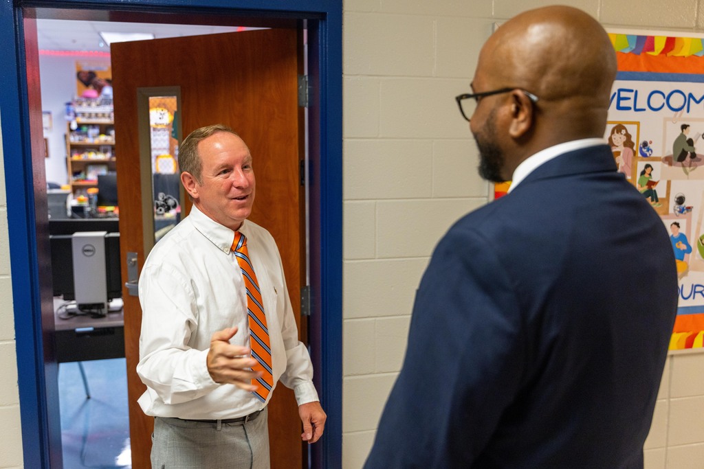 Two men in suits and ties stand in an office; one man speaks, the other listens, with a classroom door and bulletin board behind them.