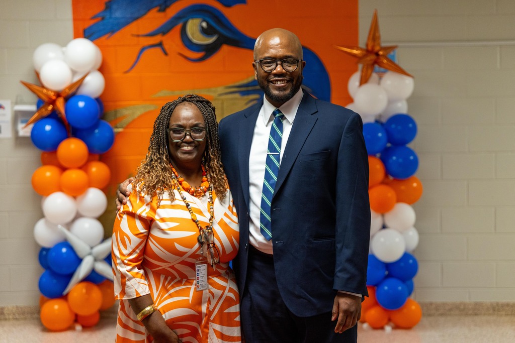A man in a blue suit and a woman in a patterned dress stand before a wall with balloons.