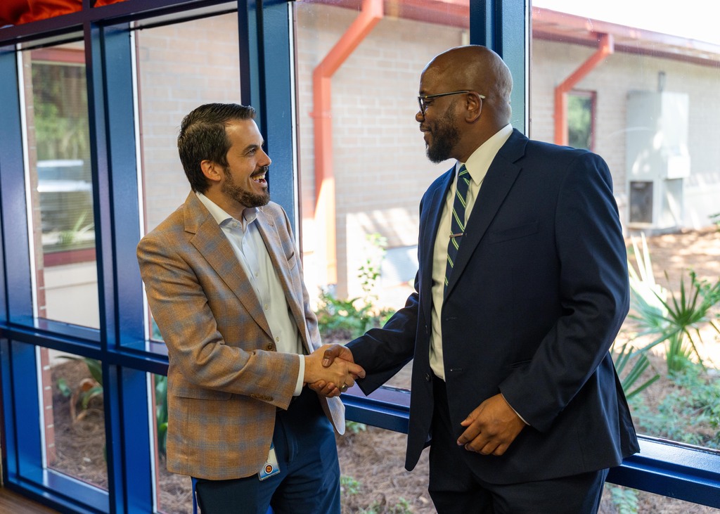 Two men in suits, one in a brown jacket and the other in a blue suit, shake hands. Behind them, a building with large windows is visible.