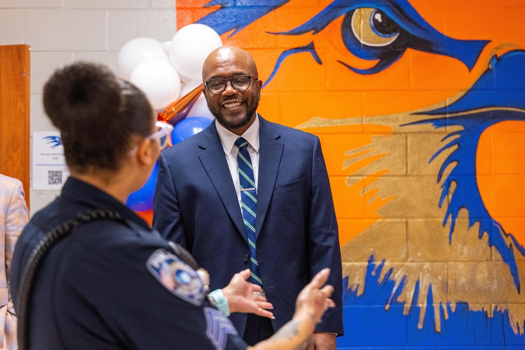 A man in a business suit and tie converses with a uniformed officer. A colorful mural of a bird is behind them.