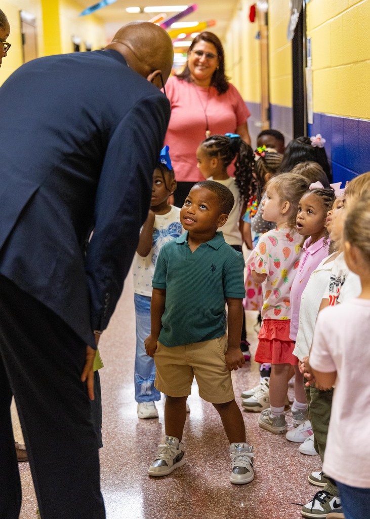 Man in suit talking to small children in hallway of school. Children wearing sneakers and some with bows.