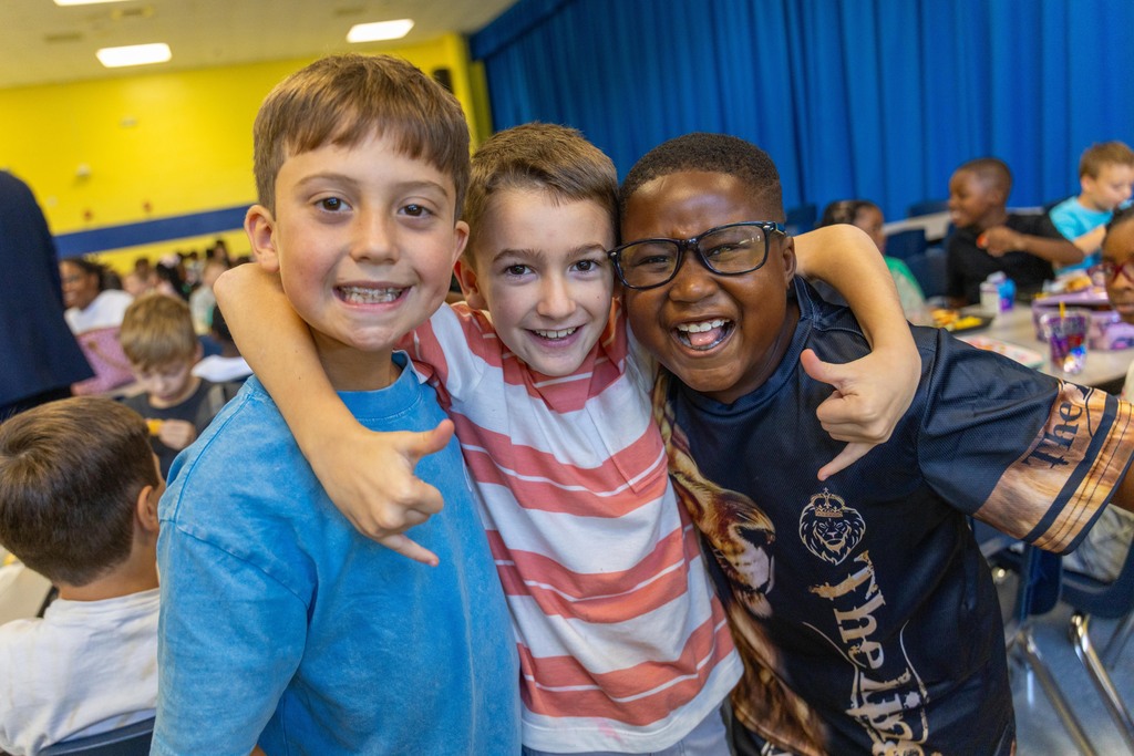 Three boys smiling, two with arms around each other, in a classroom with other children.