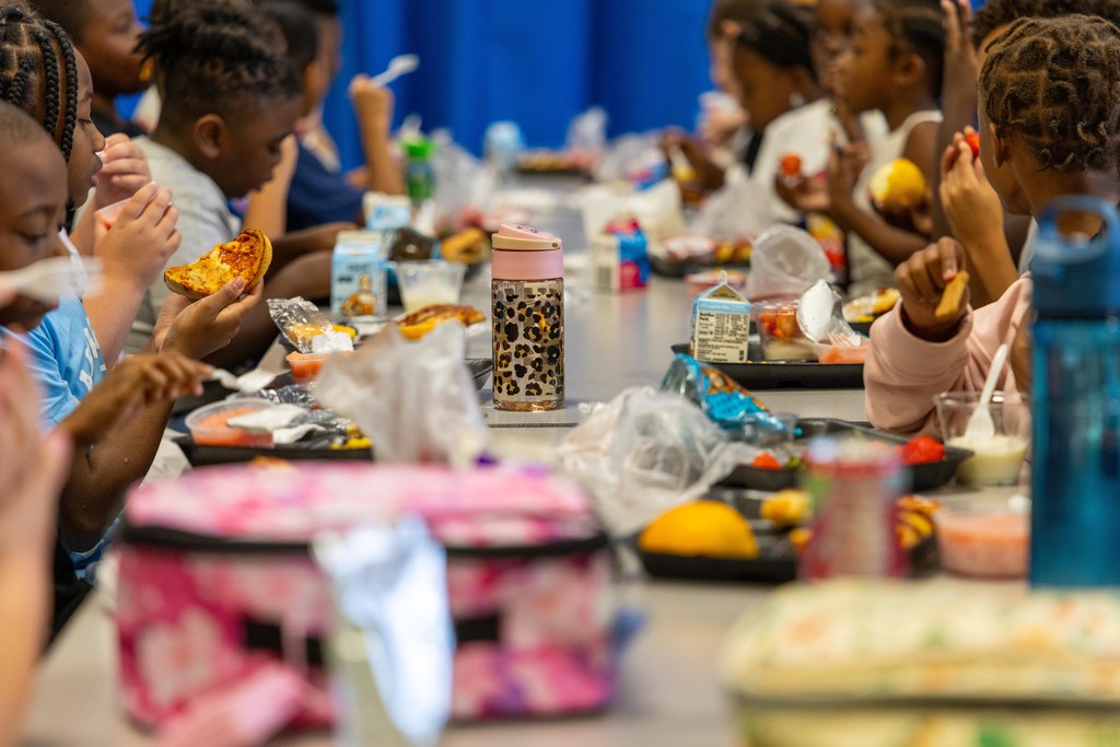 Children seated at a long table eat meals from plates, with various food items and utensils.