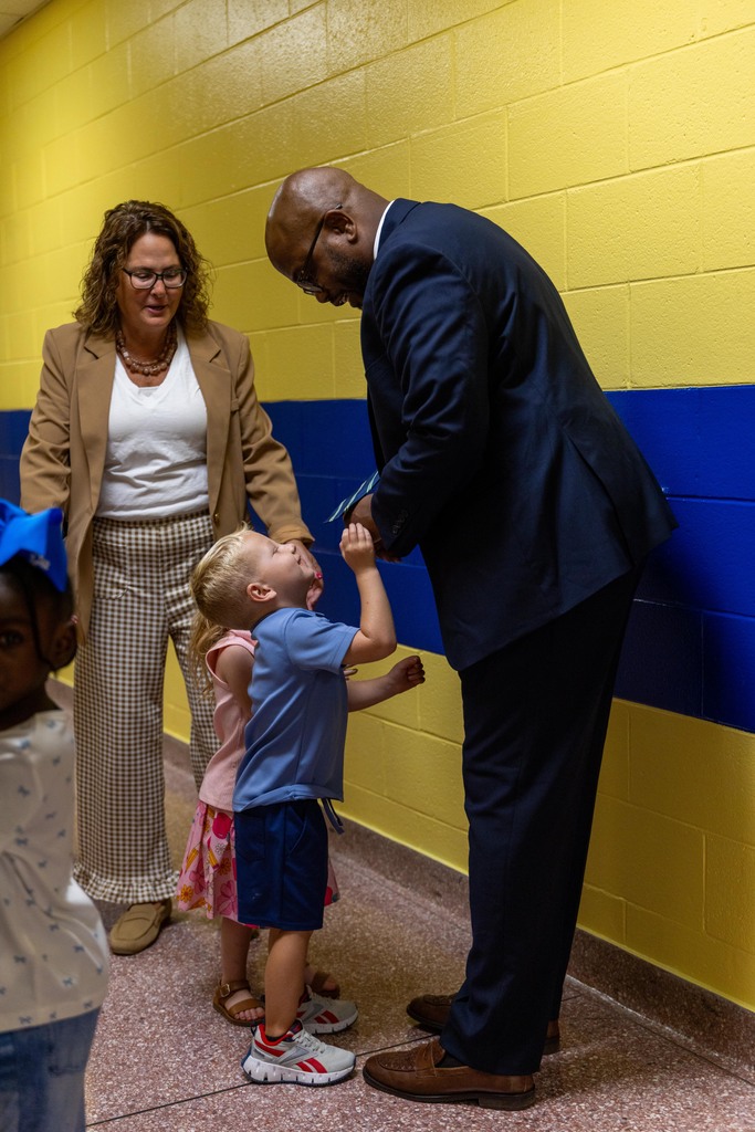 A man in a suit speaks to a young boy in a blue shirt and two girls. The boy is raising his hand. They are standing in a hallway with yellow and blue walls.