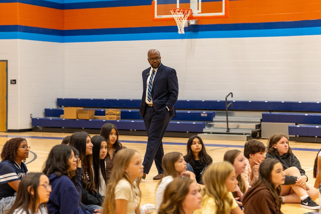 A man in a suit stands in front of a group of seated students in a gymnasium with orange and blue walls.