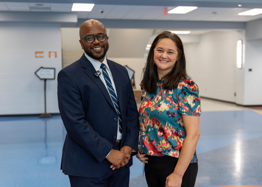 Two people stand in a hallway. A man in a suit and tie smiles, and a woman in a floral blouse smiles.