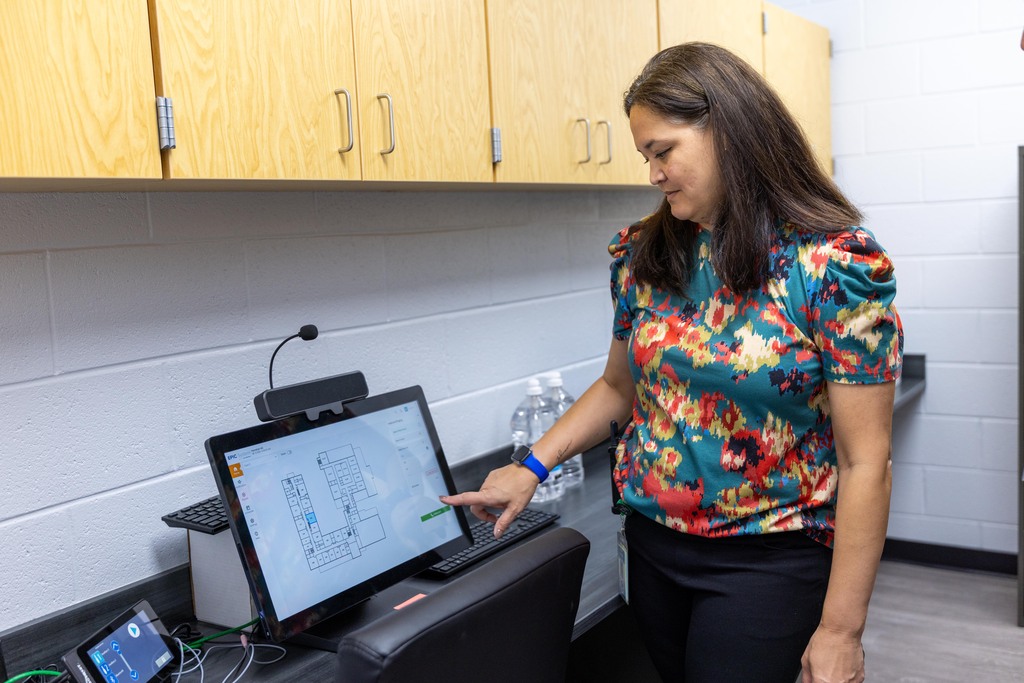 Woman in colorful shirt pointing at screen on monitor in office; cabinets, black chair, and monitor in background.