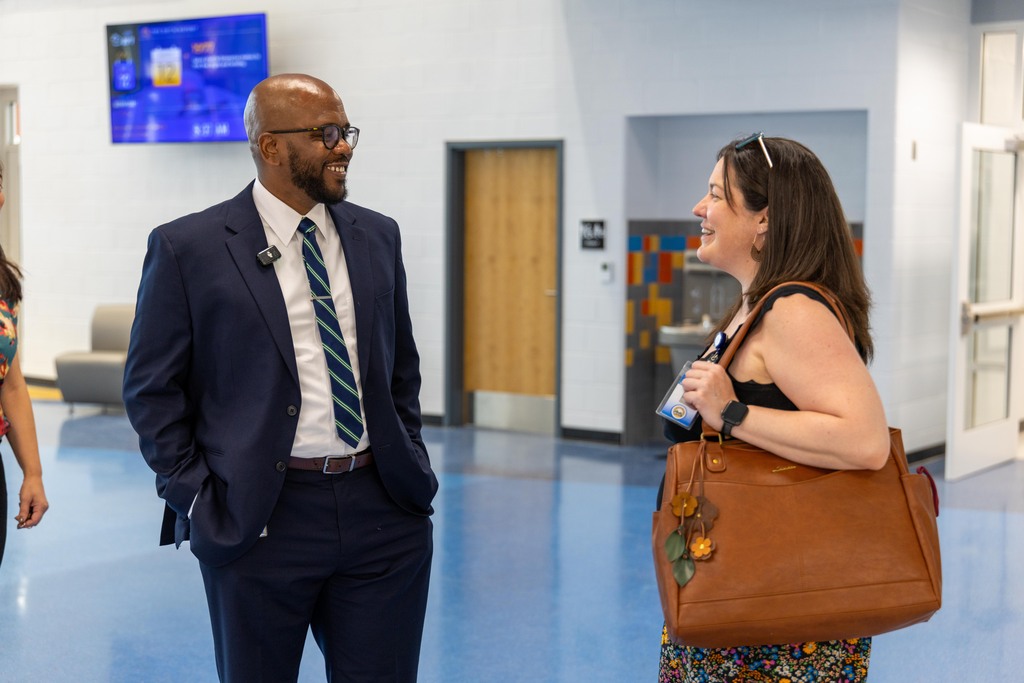 A man and a woman stand in a hallway, conversing. The man wears a suit and tie, and the woman holds a brown handbag.