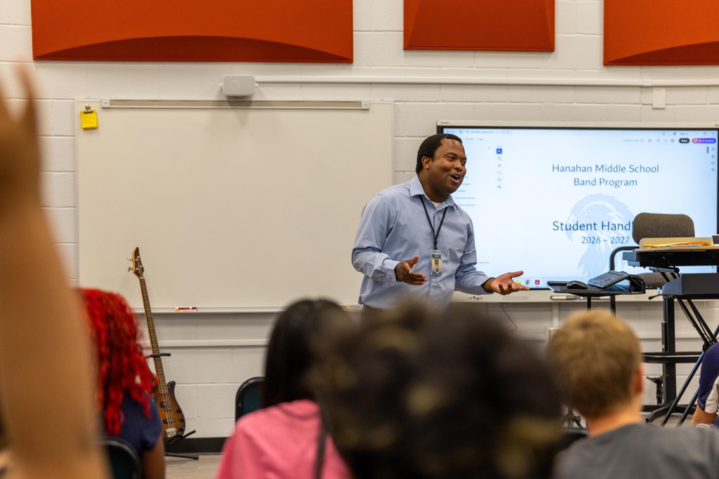 A man presents in front of a whiteboard with a projector screen behind him. Students listen and watch in a classroom.