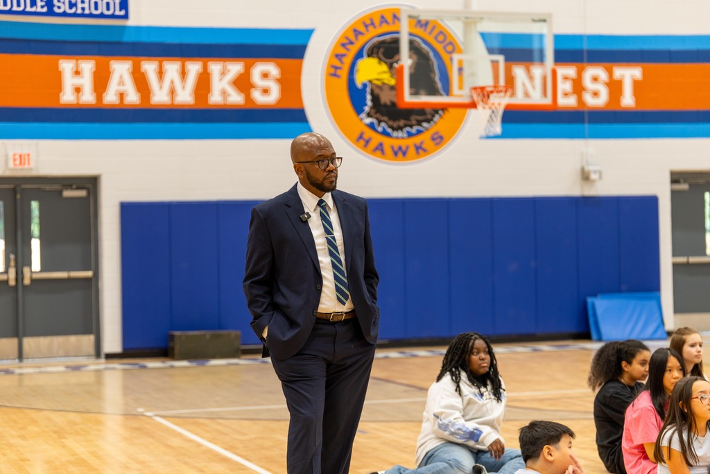 A man in a suit and tie stands in a gym with a basketball hoop. Children sit on the floor.
