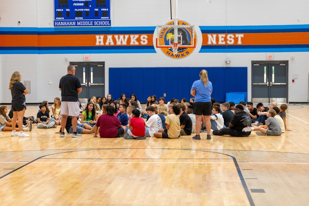 An indoor basketball court. Students sit in a circle, with two adults standing in front. A blue wall has the text "HAWKS NEST" on it.