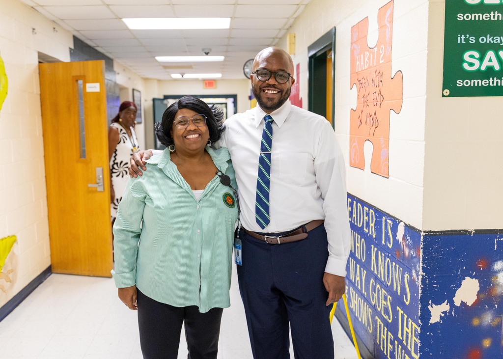 A man and woman stand in a hallway with motivational wall art, a door, and a woman in the background.