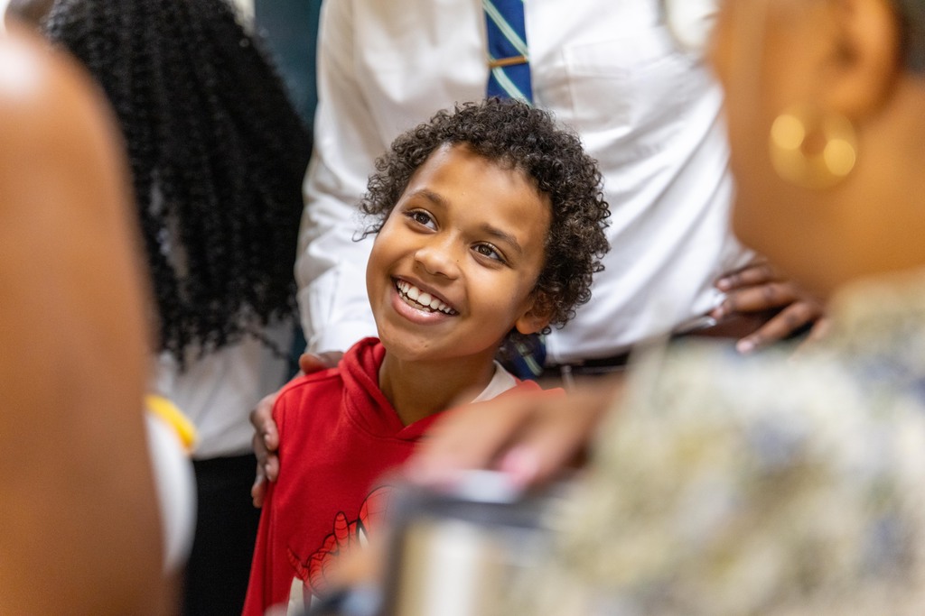 A young child with curly hair smiles, with adults standing behind, one with a hand on their shoulder.