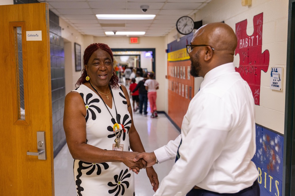 Two people shake hands in a school hallway, with a clock and posters on the wall.