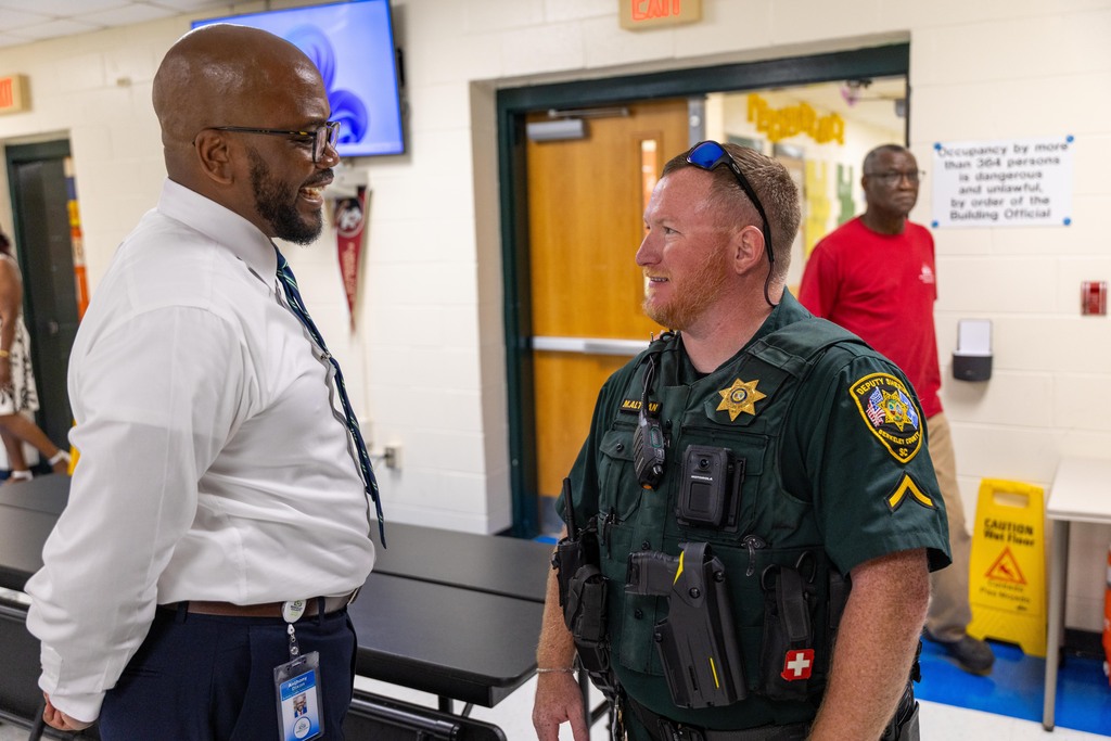 Two men speak to each other, one in a white shirt, the other in green uniform with sunglasses. Behind them, a person in a red shirt.
