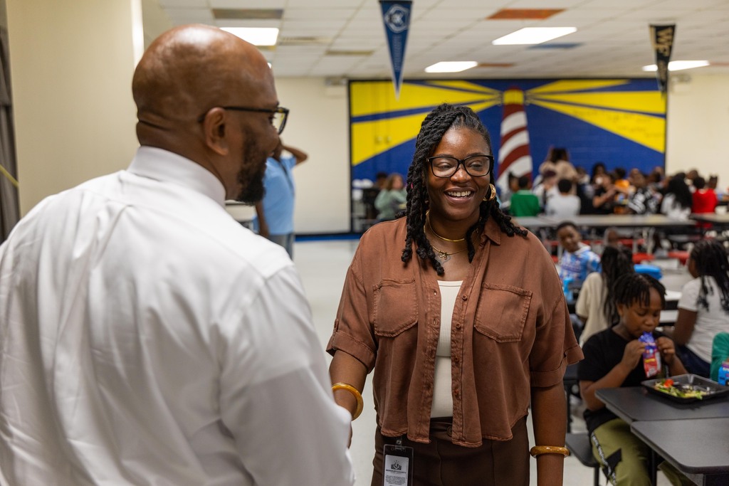 A man in a white shirt talks to a woman in a brown shirt. They are in a busy cafeteria.
