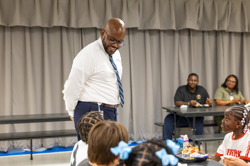 A man in a white shirt and tie stands before a group of children seated at tables.