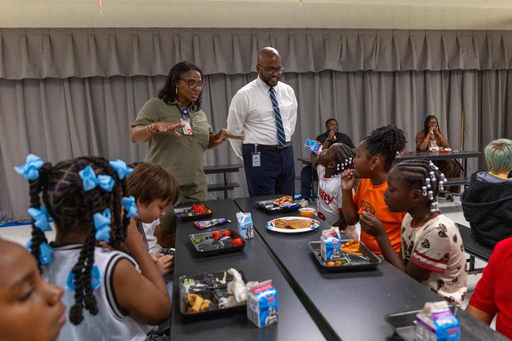 People gather in a room; a group of children sit at a table with meals. An adult woman stands next to a man in a suit.
