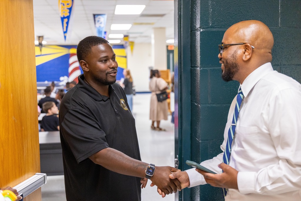 Two men in a school hallway, one in black with a watch, the other in white with a tie, shaking hands.