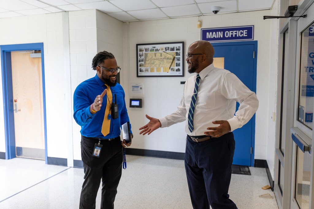 Two men in suits, one holding a folder, talk in an office hallway with a blue door and a picture frame.