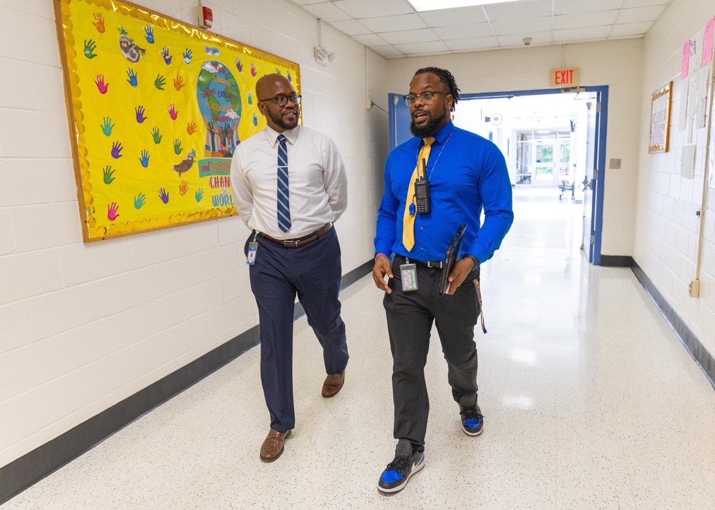 Two men in formal attire, one in blue and one in white, walking down a brightly lit hallway.