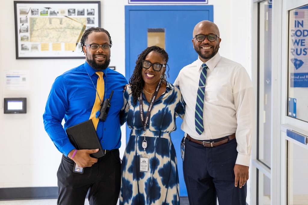 Three people in formal attire stand near a blue door. Two men and one woman. One man holds a folder.