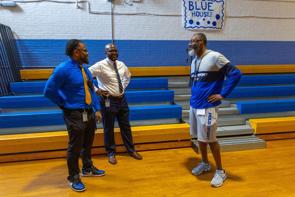 Three men stand in a gymnasium, with blue and yellow bleachers behind them. A sign reading "Blue House" is mounted on the wall.