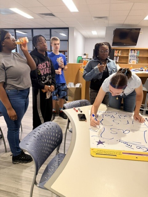 Five people standing in a room, one person leaning over a table with a large sheet of paper.