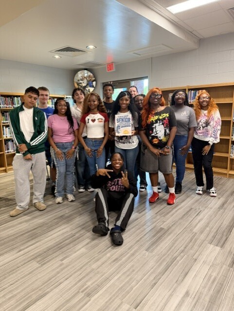 A group of students pose for a photo in a library with wooden shelves and a wooden floor.