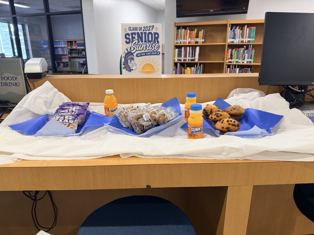 Display table with cookies, muffins, and orange juice. Behind it, a bookshelf with books and a banner.