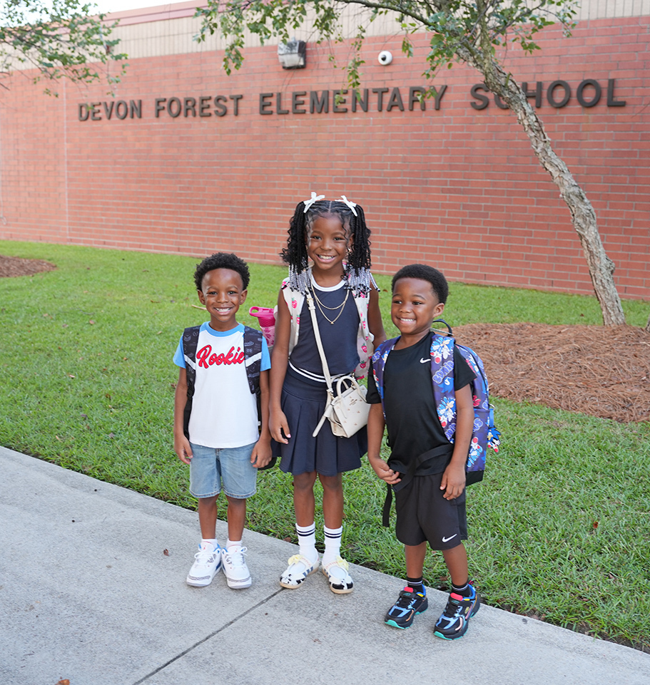 Three children stand on a sidewalk, two boys and a girl, in front of a brick building.