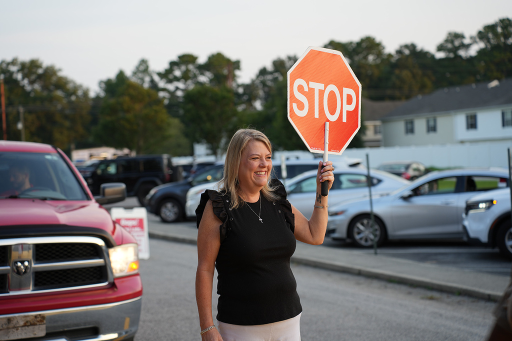 A woman stands in a parking lot holding a stop sign, with cars parked and trees in the background.