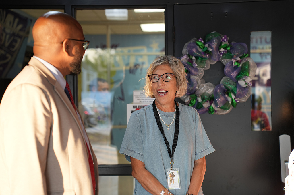 Man in glasses and suit talks to a smiling woman in glasses, blue shirt, and lanyard. Behind them is a door with decorative wreath.