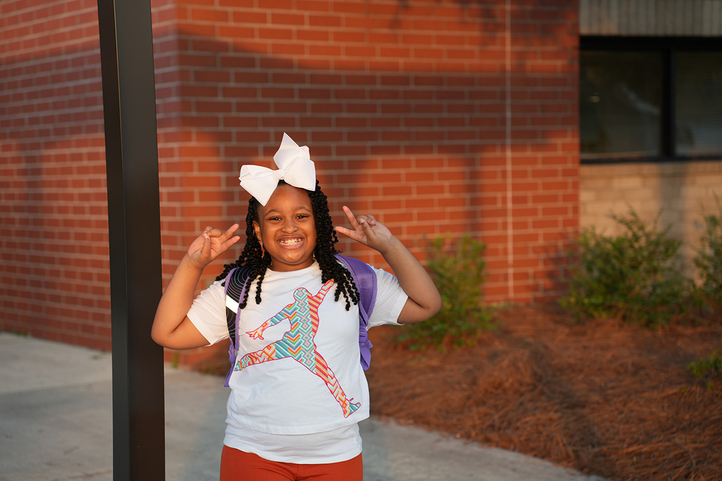 A girl with a white bow, wearing a white shirt and red pants, stands outside a brick building.