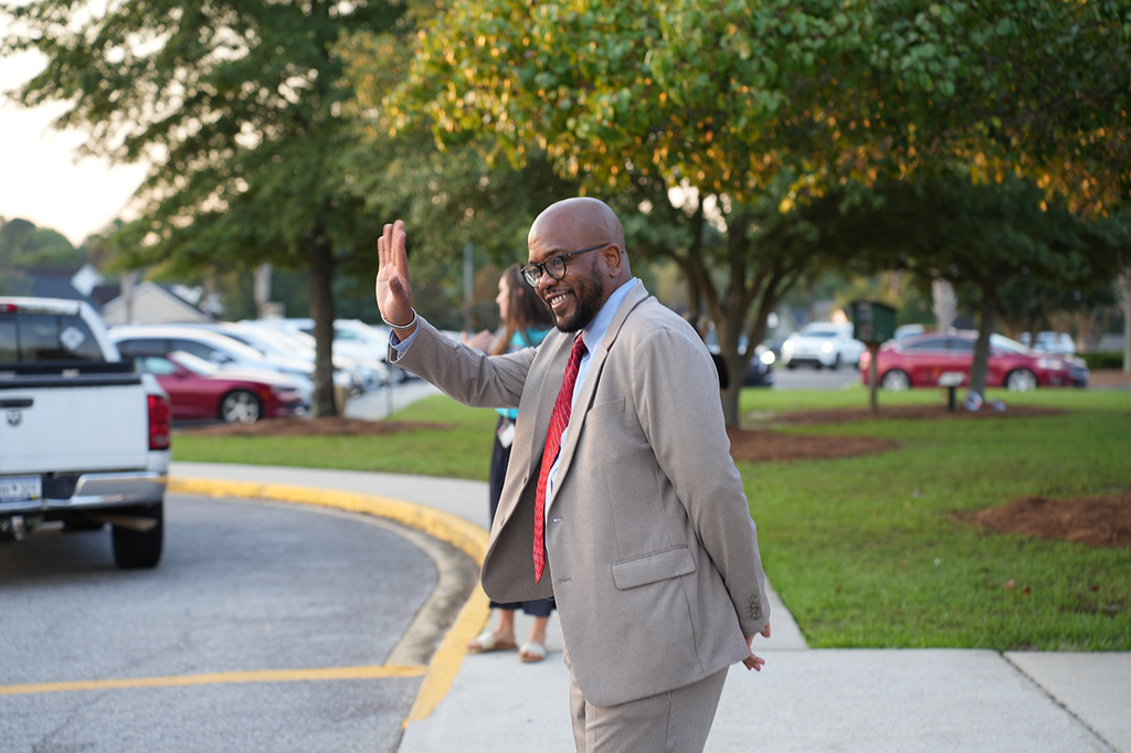 A man in a tan suit waves to the camera, standing in a parking lot with green grass and trees.