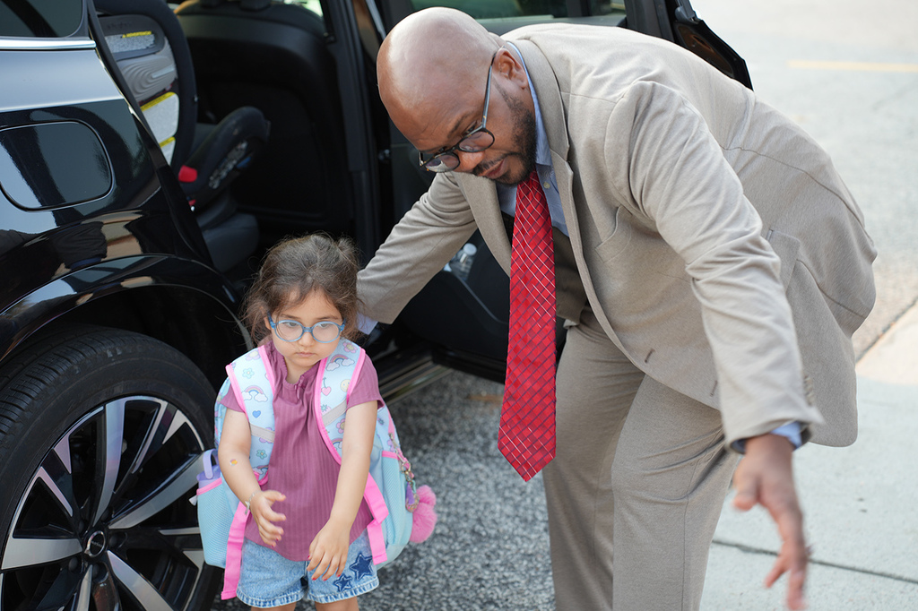 A man helps a young child with a backpack exit a car in a parking lot.