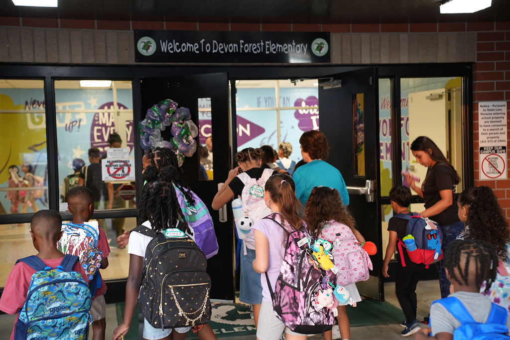 Children and adults with backpacks at a school entrance. A sign reads "Welcome to Devon Forest Elementary."