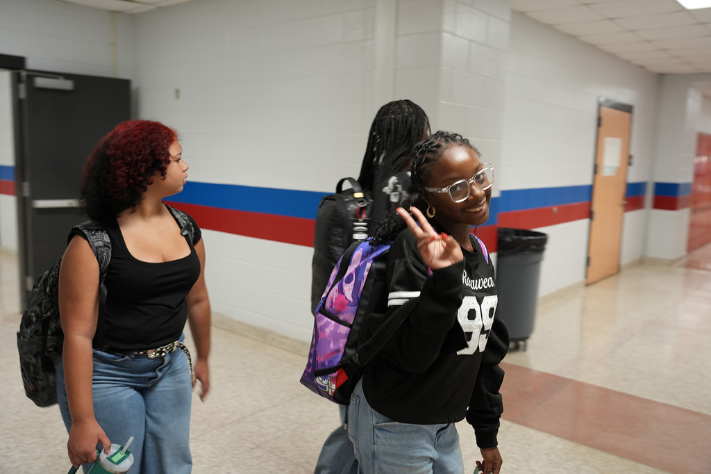 Three women walking in a hallway; one in front has curly hair, wears a black tank top and jeans.