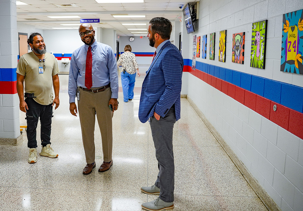 Three men in a hallway. Two men in business attire, one in a red tie, the other in a blue jacket.