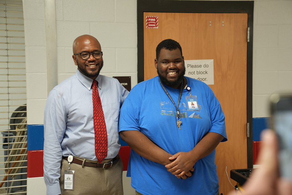 Two men stand in front of a door with a sign. One wears a blue shirt, the other a blue shirt and red tie.