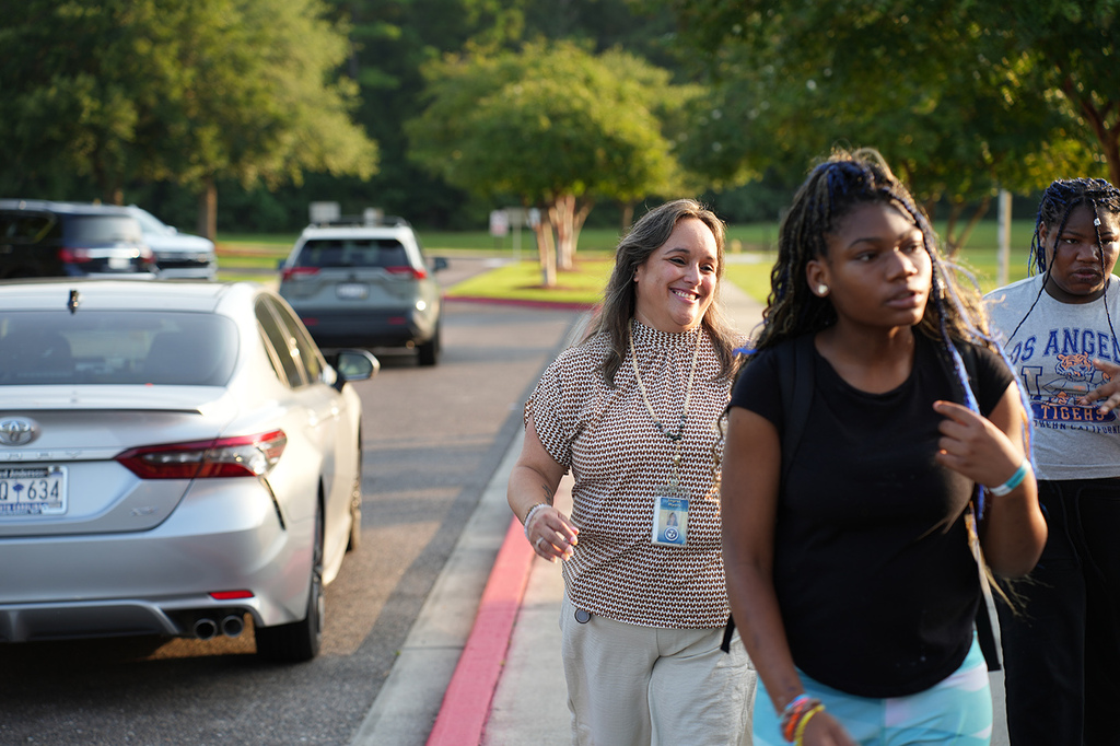 People walking on a sidewalk next to a street. One woman holds a paper, and another wears a bracelet. Cars are parked.