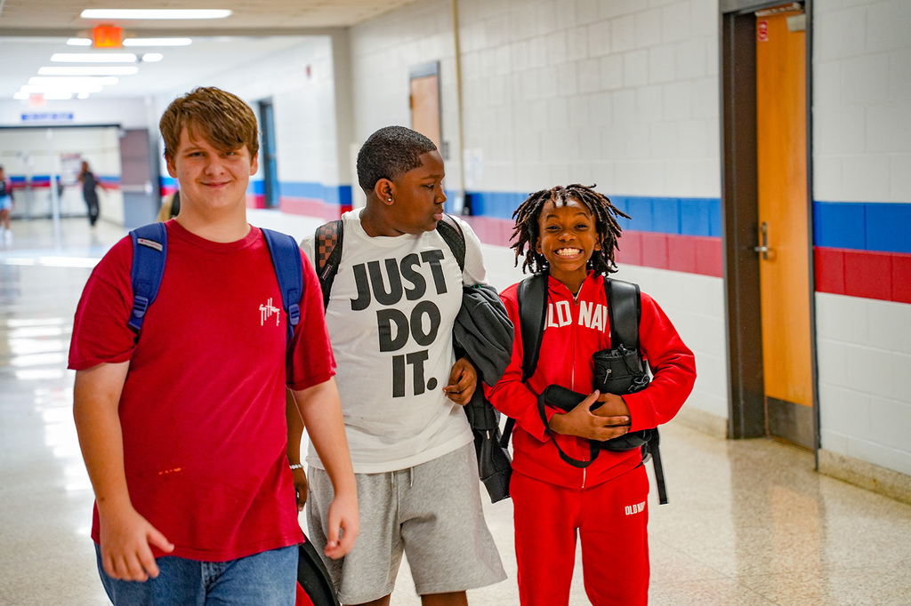 Three students walk in a school hallway, each with a backpack. One wears a red shirt, another a red jumpsuit, and the middle one a white shirt.