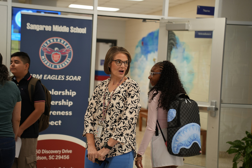 Several students and an adult woman with glasses stand by a colorful banner and door in a hallway.