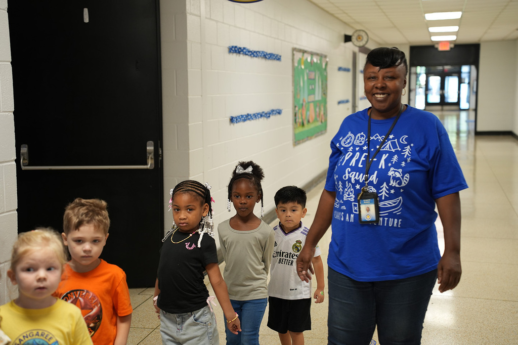 A woman in a blue shirt guides a line of children down a school hallway, past colorful posters.